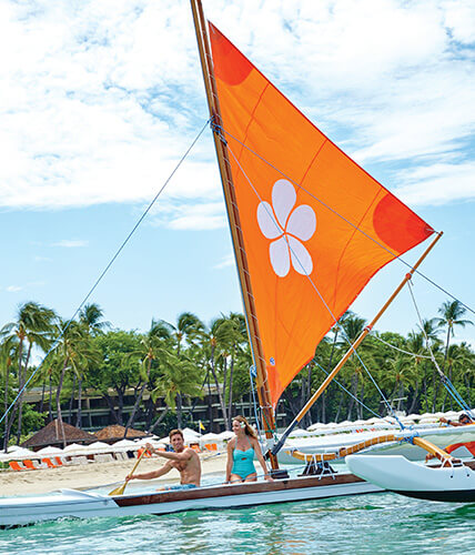 A young man and woman paddle an outrigger canoe with an orange sail featuring a white plumeria blossom design.